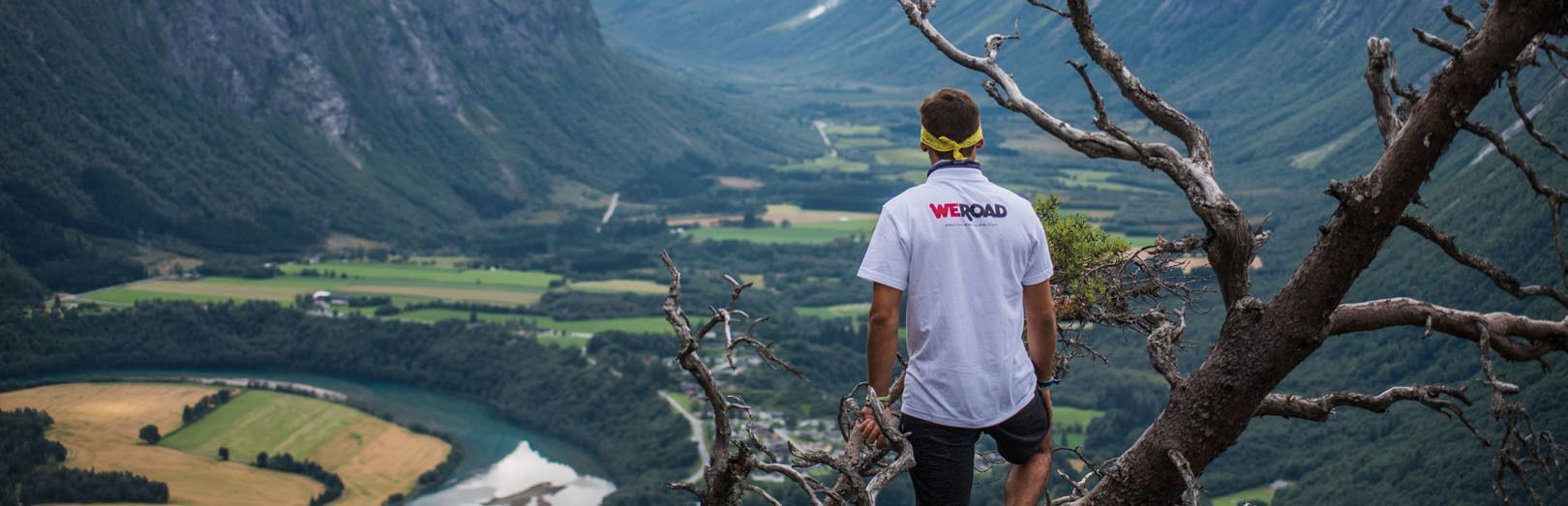 Un hombre con una camiseta de WeRoad de pie en un acantilado, contemplando un vasto valle verde con un río y montañas.