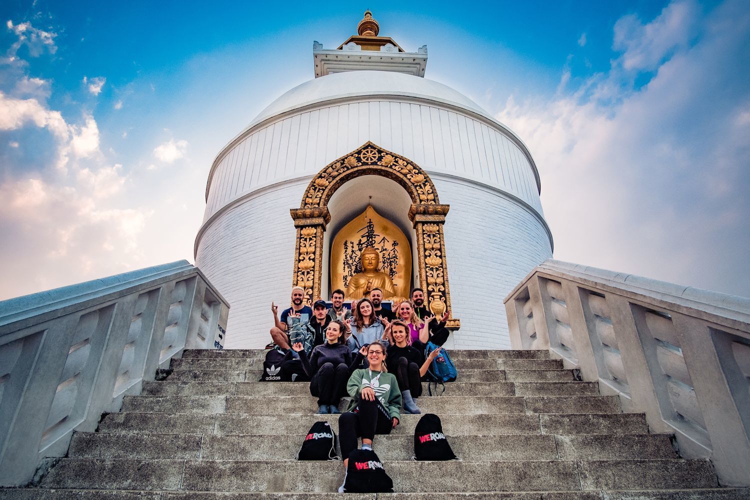 Un groupe WeRoad pose pour une photo sur les marches d'un stupa blanc, avec une statue de Bouddha dorée dans une niche derrière eux.