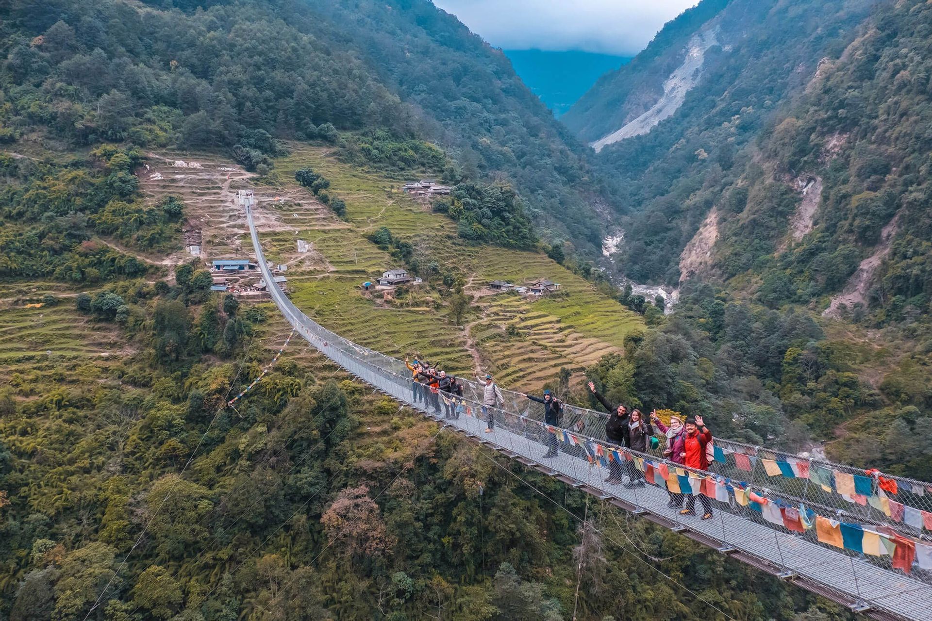 Un gruppo WeRoad saluta da un lungo ponte sospeso, decorato con bandiere colorate, che attraversa una profonda valle con montagne verdi e terrazzate.