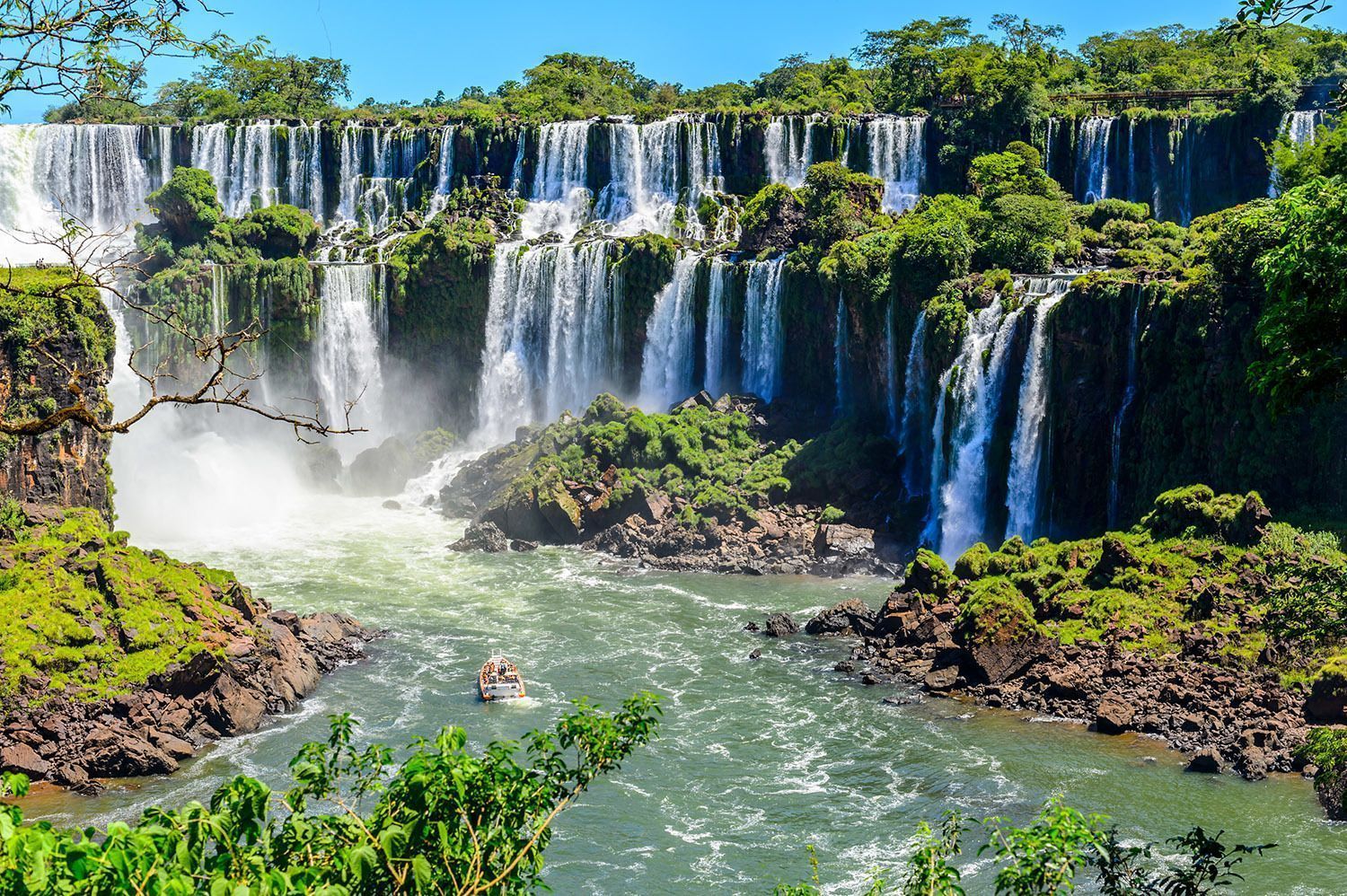 A small tour boat navigates the turbulent river at the base of a wide, multi-tiered waterfall covered in lush green vegetation.