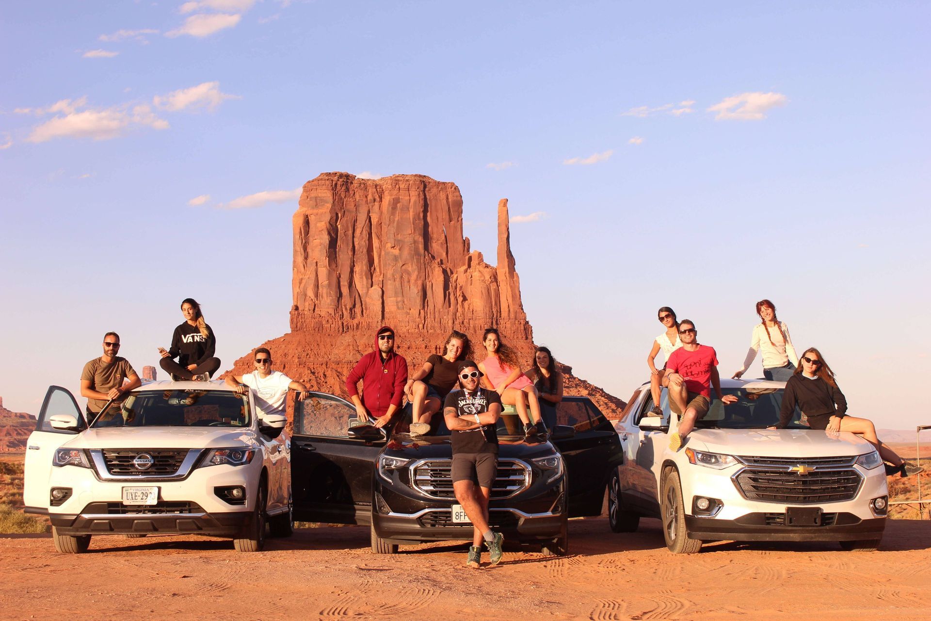A WeRoad group trip poses on and around three SUVs in a desert with towering red rock formations in the background.