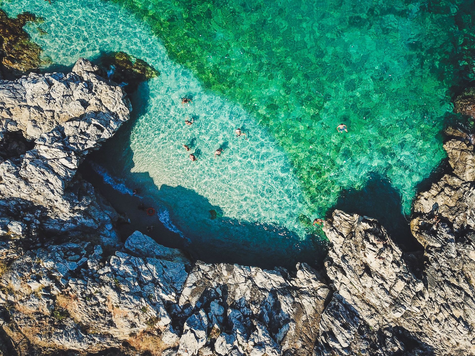 Una vista aerea di persone che nuotano nelle acque limpide e turchesi di una cala rocciosa.