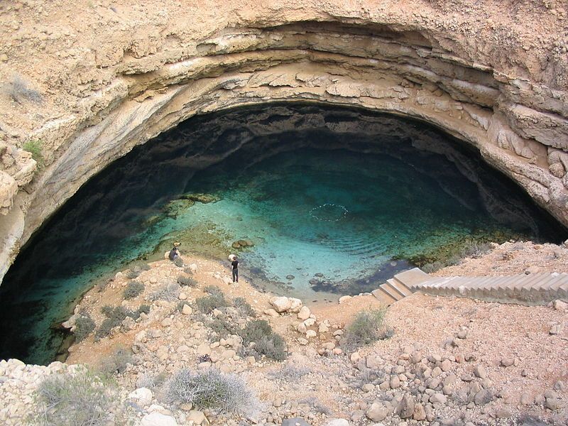 Un'ampia veduta di un grande cenote circolare pieno d'acqua turchese, con due persone in piedi sul bordo roccioso.