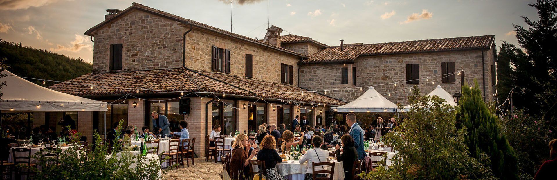 Un folto gruppo di persone che cena ai tavoli su un patio in pietra di fronte a una villa rustica, con lucine a catena sopra al crepuscolo.
