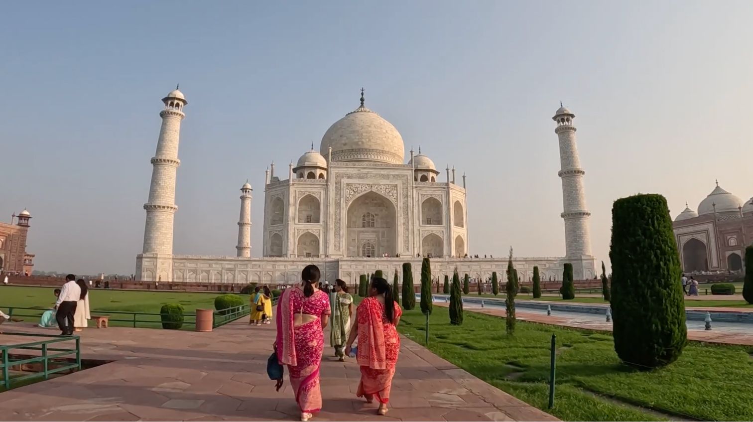 Two women in colorful saris walk on a stone pathway towards a large, white marble mausoleum with manicured gardens.