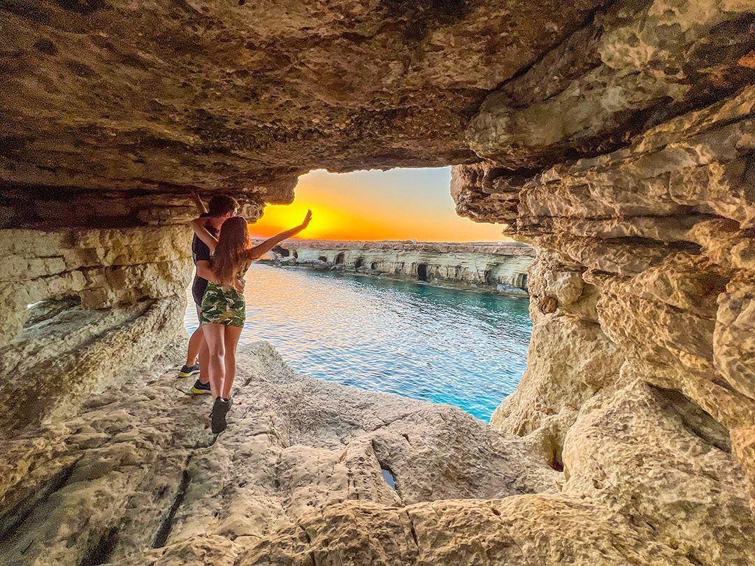 Una coppia si abbraccia all'interno di una grotta marina rocciosa, guardando il tramonto su un mare calmo.