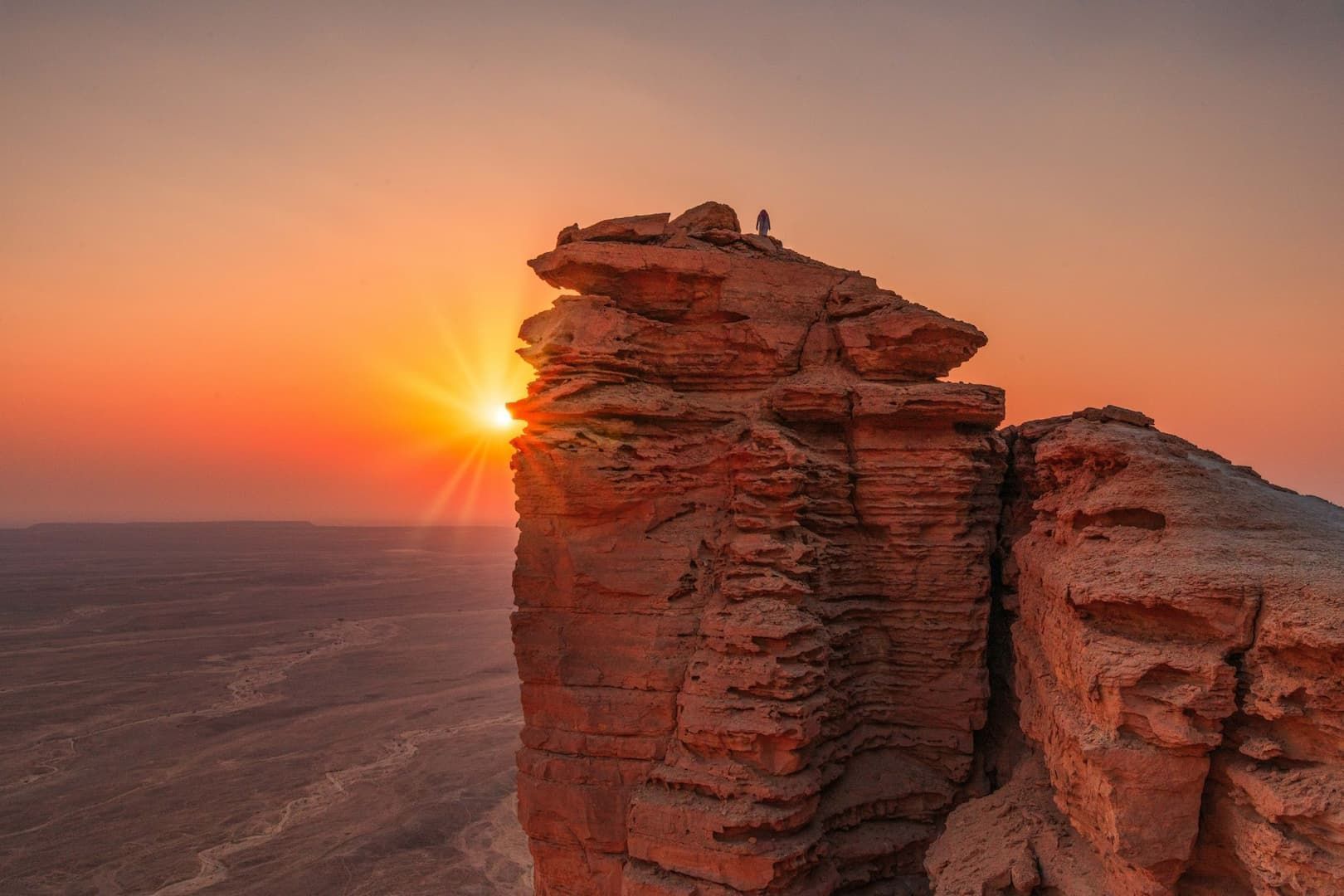 Una persona si trova su un'imponente formazione rocciosa che domina un vasto paesaggio desertico durante un vivace tramonto arancione.