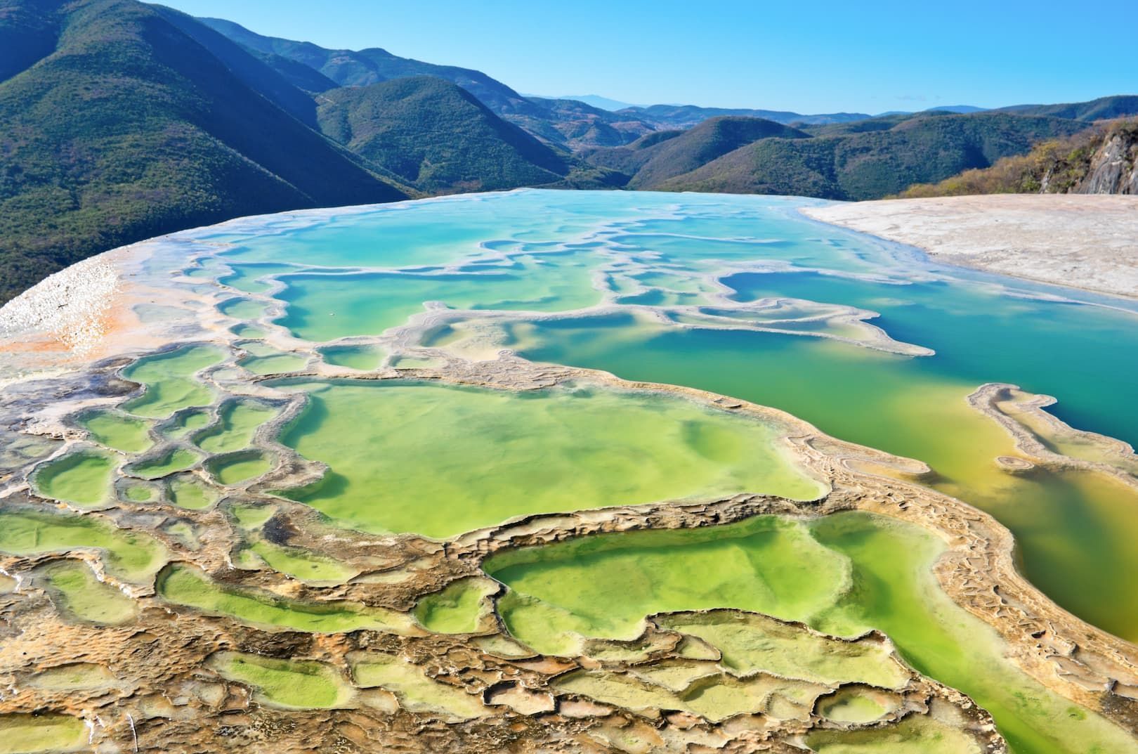 Piscinas minerales en terrazas de verde y turquesa vibrante, con vistas a una vasta cordillera bajo un cielo azul claro.