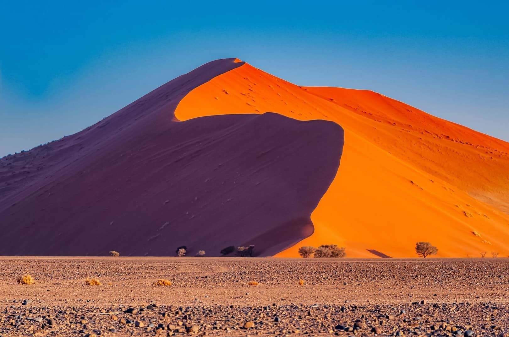 Una gran duna de arena naranja proyecta una sombra profunda sobre sí misma en un paisaje desértico rocoso bajo un cielo azul claro.