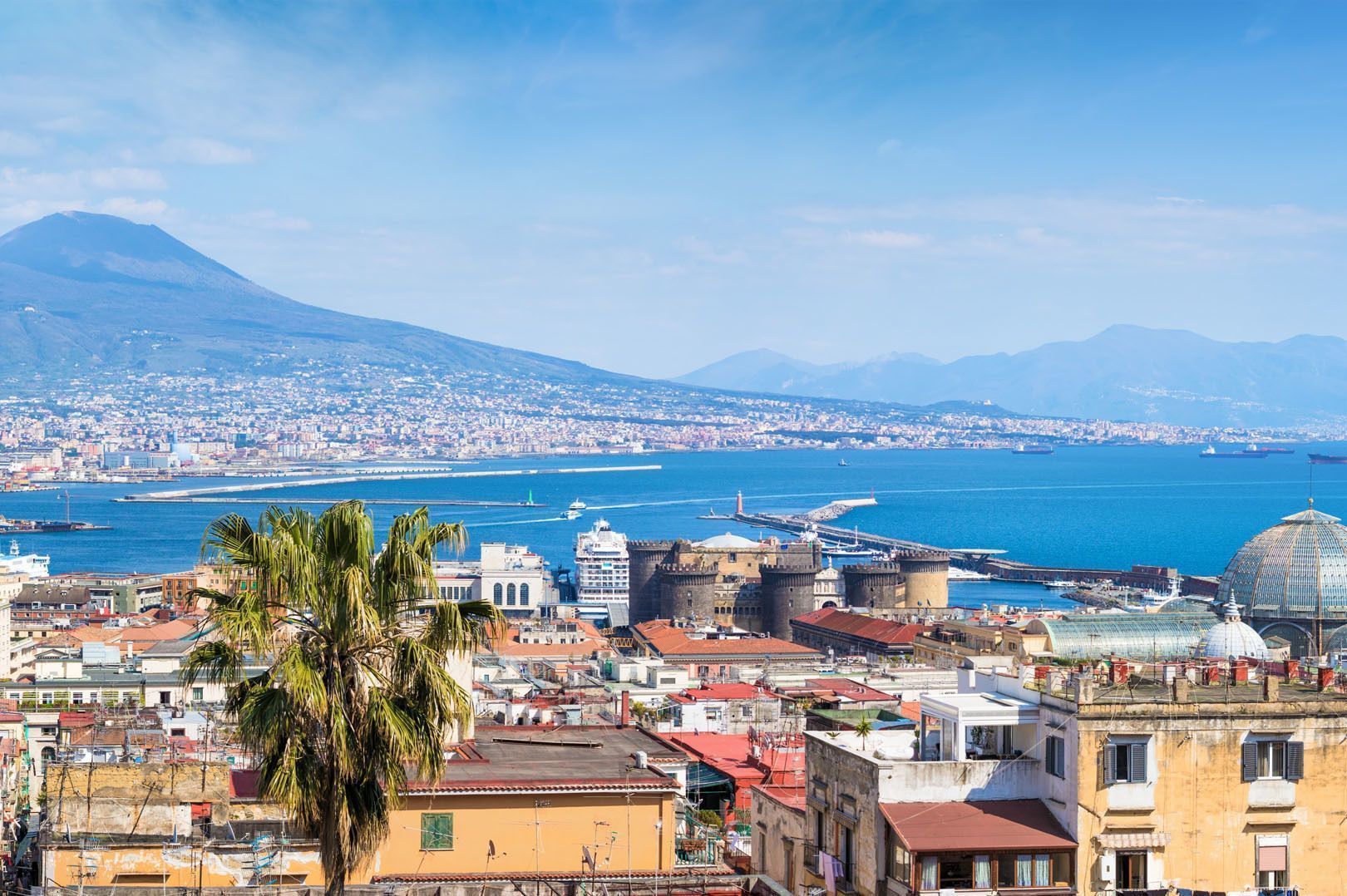Vista dall'alto di una città costiera con un grande vulcano sullo sfondo, che mostra tetti, una palma e una baia blu brillante.