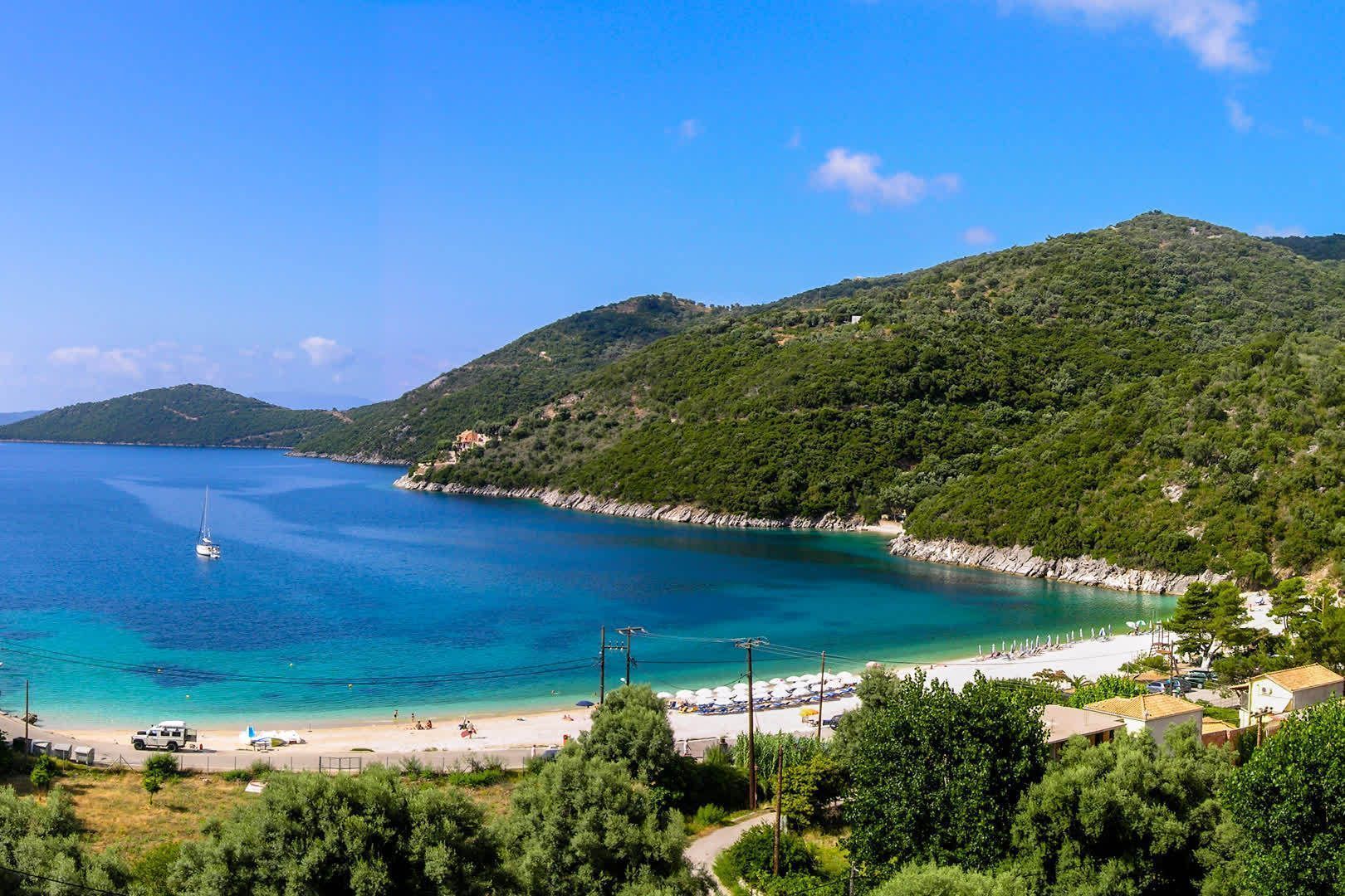 Ein Panoramablick auf eine türkisfarbene Bucht mit einem weißen Sandstrand und einem einsamen Segelboot, umrahmt von bewaldeten Hügeln.