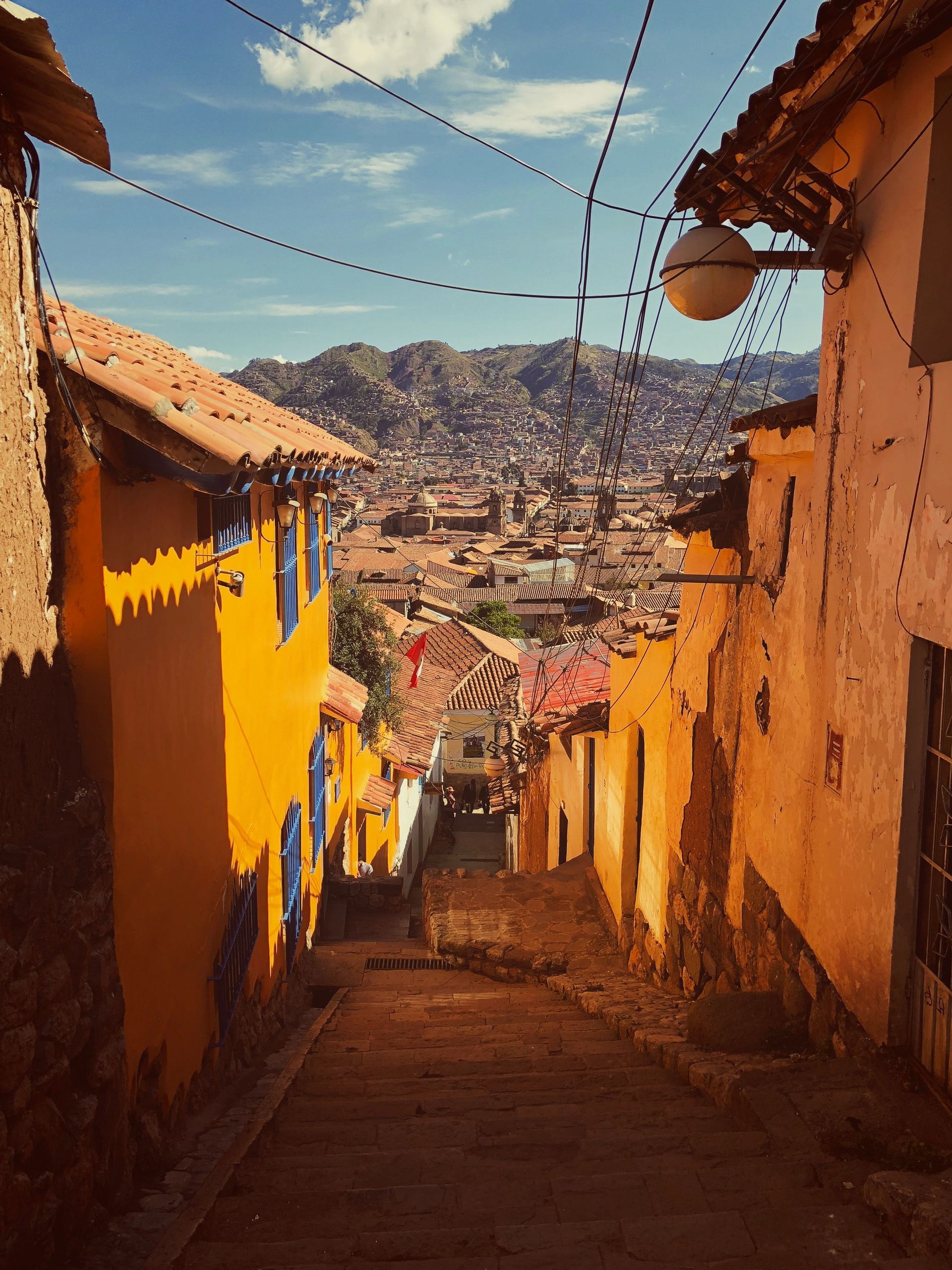 Un callejón de piedra estrecho y soleado, con escaleras, desciende entre edificios coloridos, revelando una vista de la ciudad y las montañas.