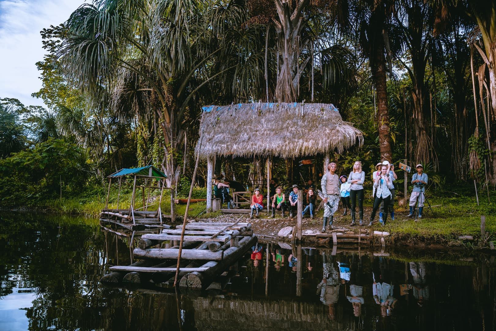 Un viaje en grupo de WeRoad posa en la orilla de un río en la jungla junto a un muelle de madera y una cabaña con techo de paja, rodeados de palmeras.