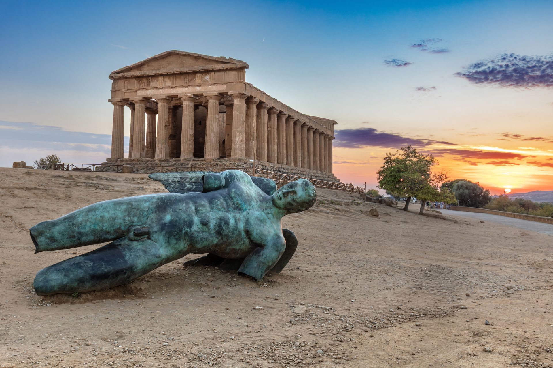 Una statua di bronzo di una figura alata caduta giace su una collina polverosa con un antico tempio greco sullo sfondo al tramonto.