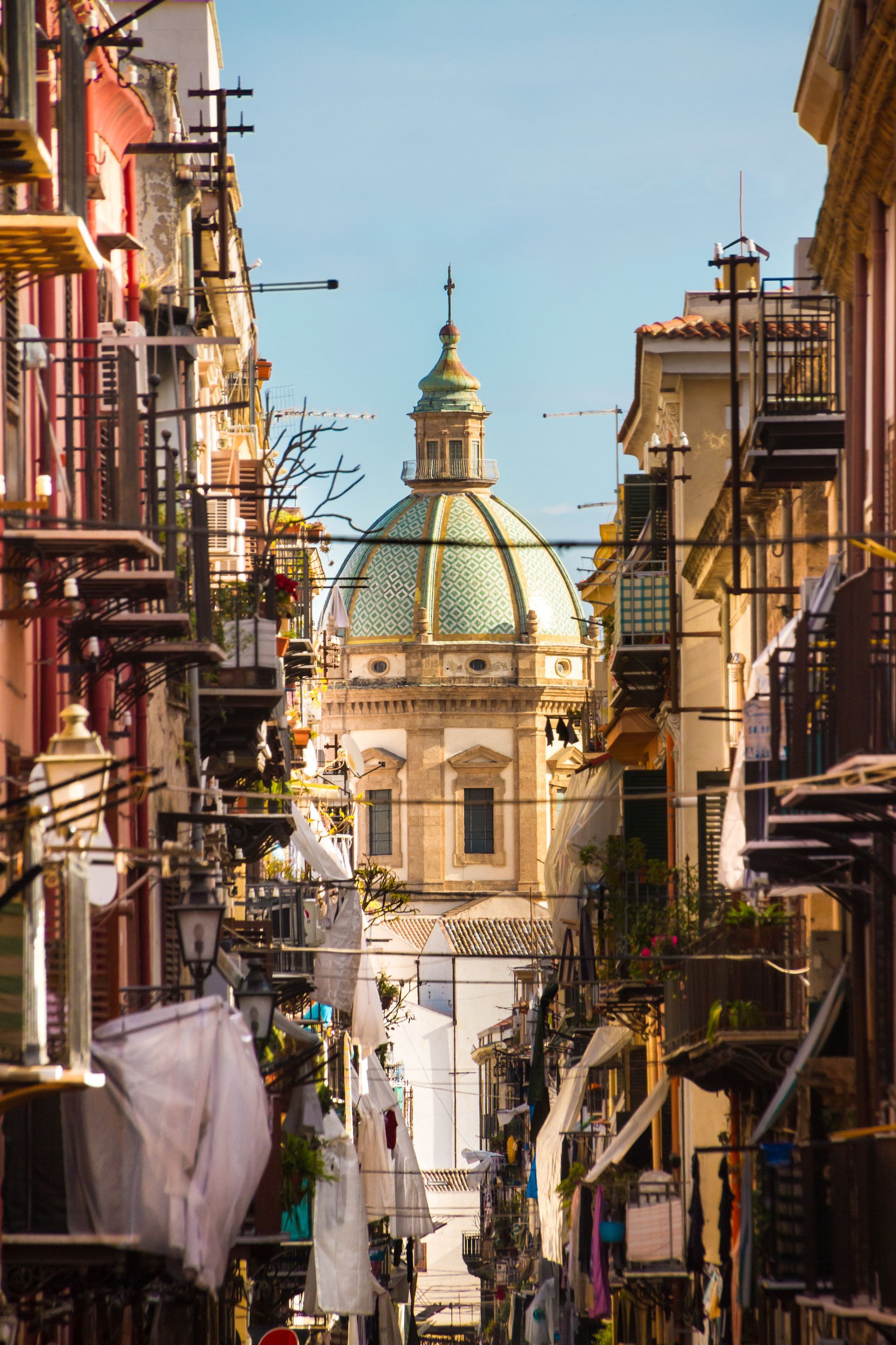 Una cupola dalle tegole verdi di una chiesa è visibile alla fine di una via stretta, fiancheggiata da edifici colorati e panni stesi.