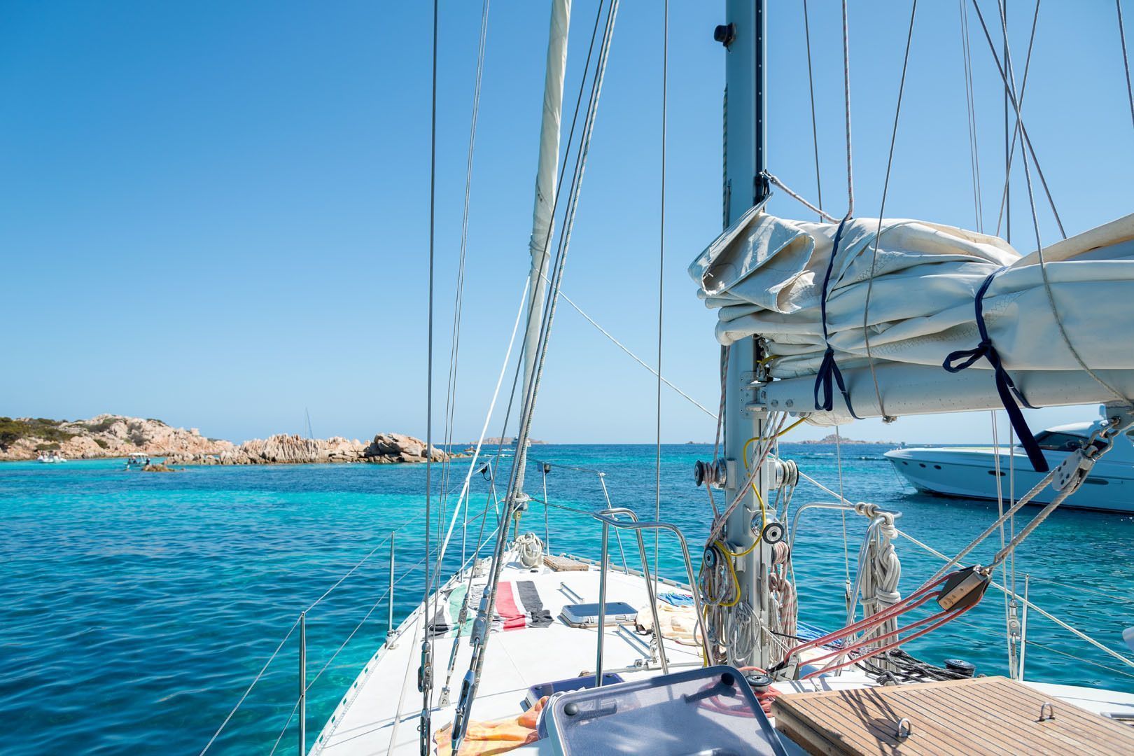 Vista dal ponte di una barca a vela su acqua cristallina turchese verso una costa rocciosa sotto un cielo azzurro brillante.