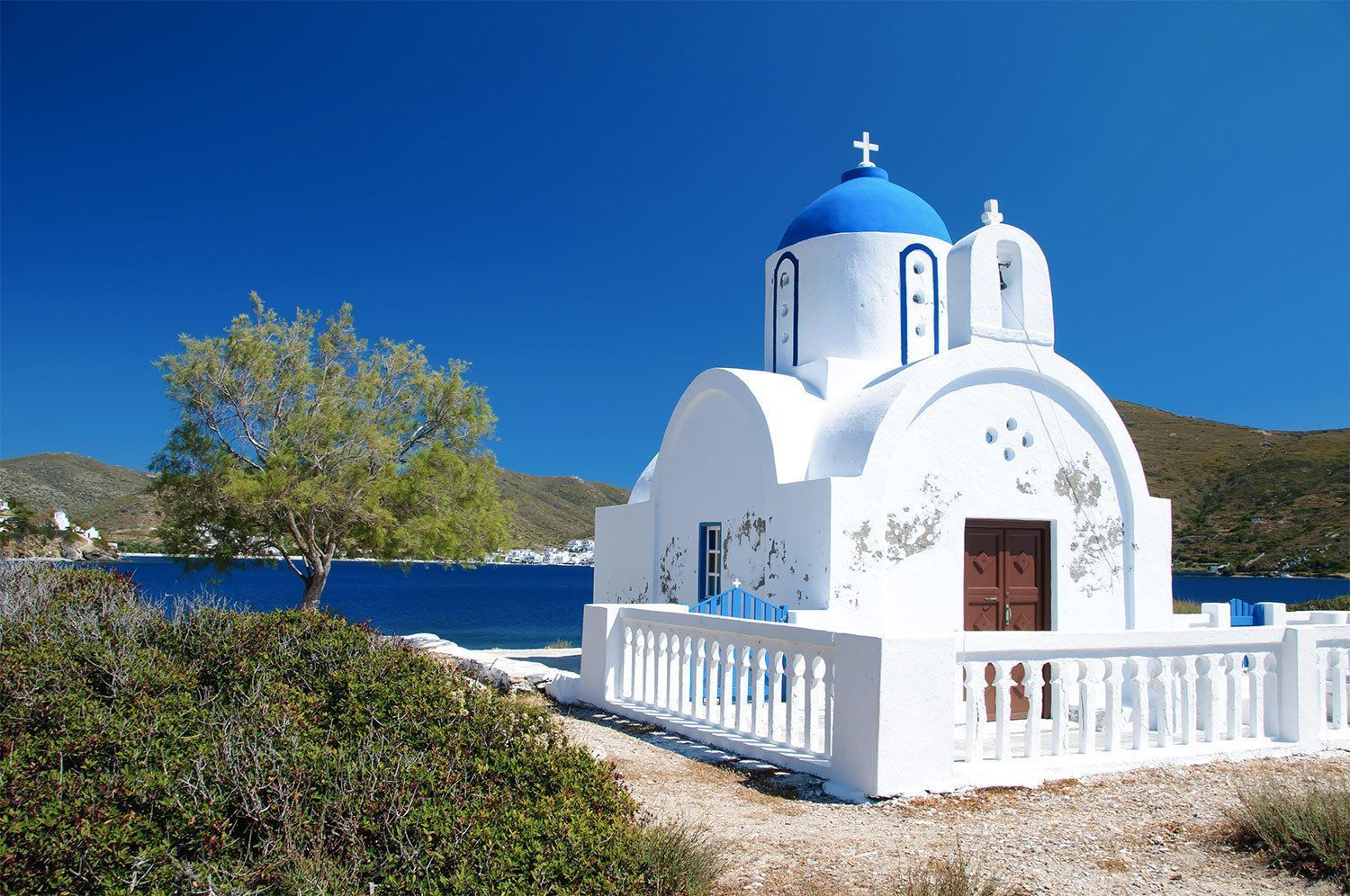 Una chiesa bianca con una cupola blu si erge vicino alla costa affacciata sul mare sotto un cielo azzurro.