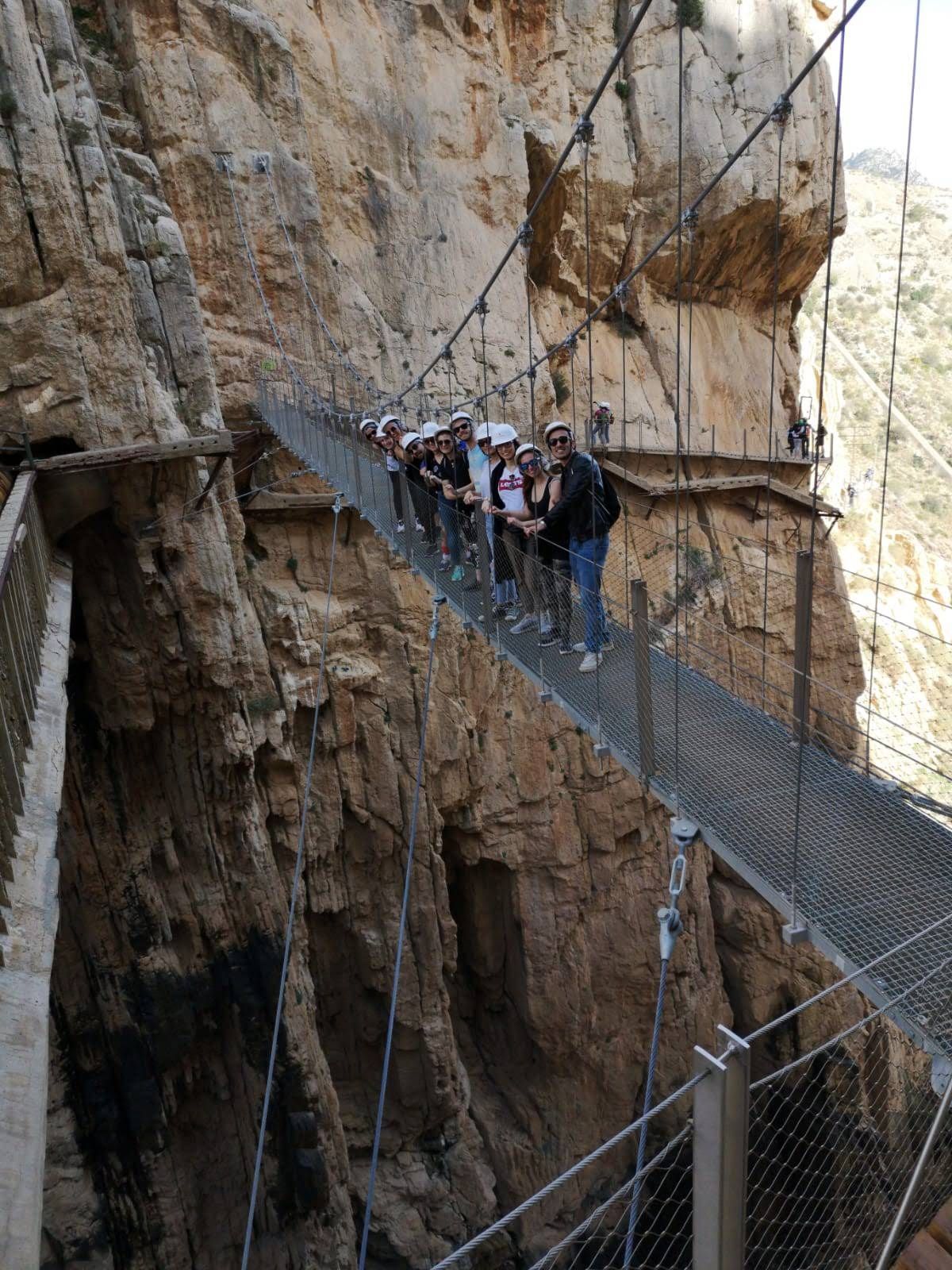 Eine WeRoad-Reisegruppe mit Helmen posiert für ein Foto auf einer Hängebrücke, die an einer steilen Canyonwand befestigt ist.