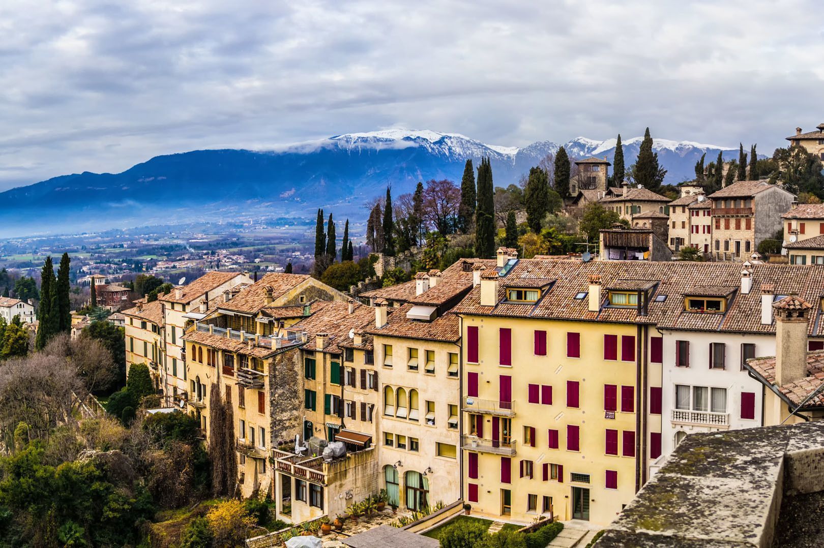 Un borgo collinare con edifici dai tetti in terracotta, annidato ai piedi di una grande catena montuosa innevata, sotto un cielo nuvoloso.