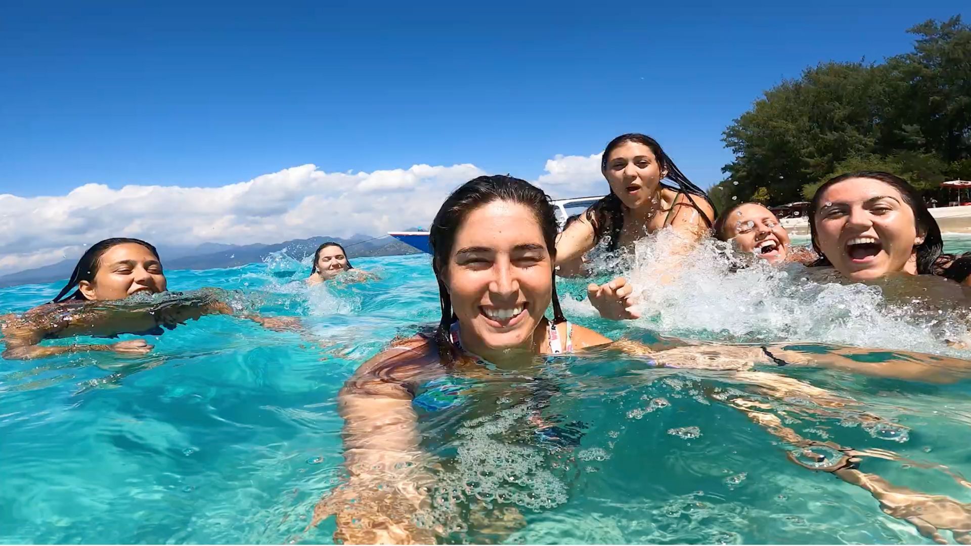 Un viaje en grupo de WeRoad con mujeres sonrientes nadando y chapoteando en agua turquesa en un día soleado cerca de una playa.