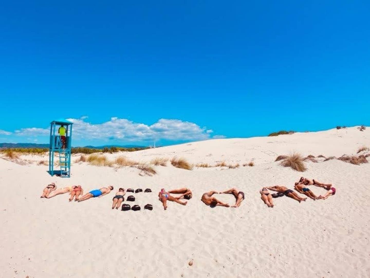 Un groupe WeRoad en voyage, étendu sur une plage de sable blanc, formant le mot WEROAD avec leurs corps et leurs sacs sous un ciel bleu.
