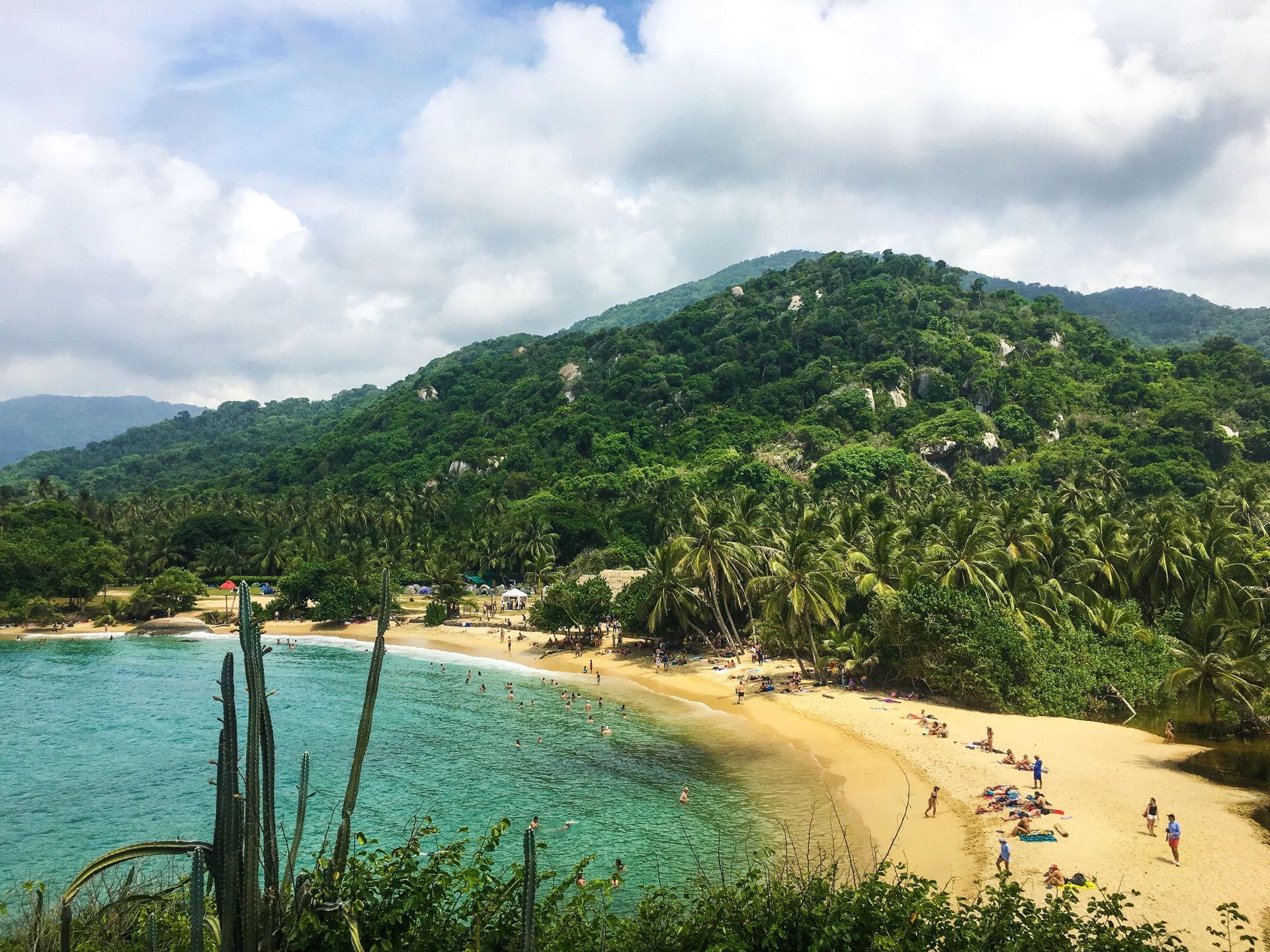A high-angle view of a tropical cove with a sandy beach, turquoise water, and a lush green hill covered in forest.