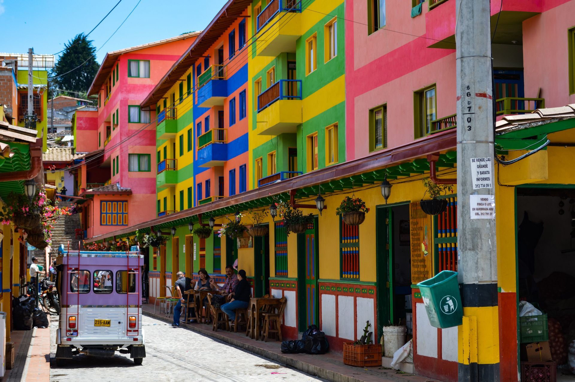 Una calle empedrada bordeada de edificios a rayas de colores vivos, donde un vehículo morado está estacionado y la gente se sienta en mesas al aire libre.
