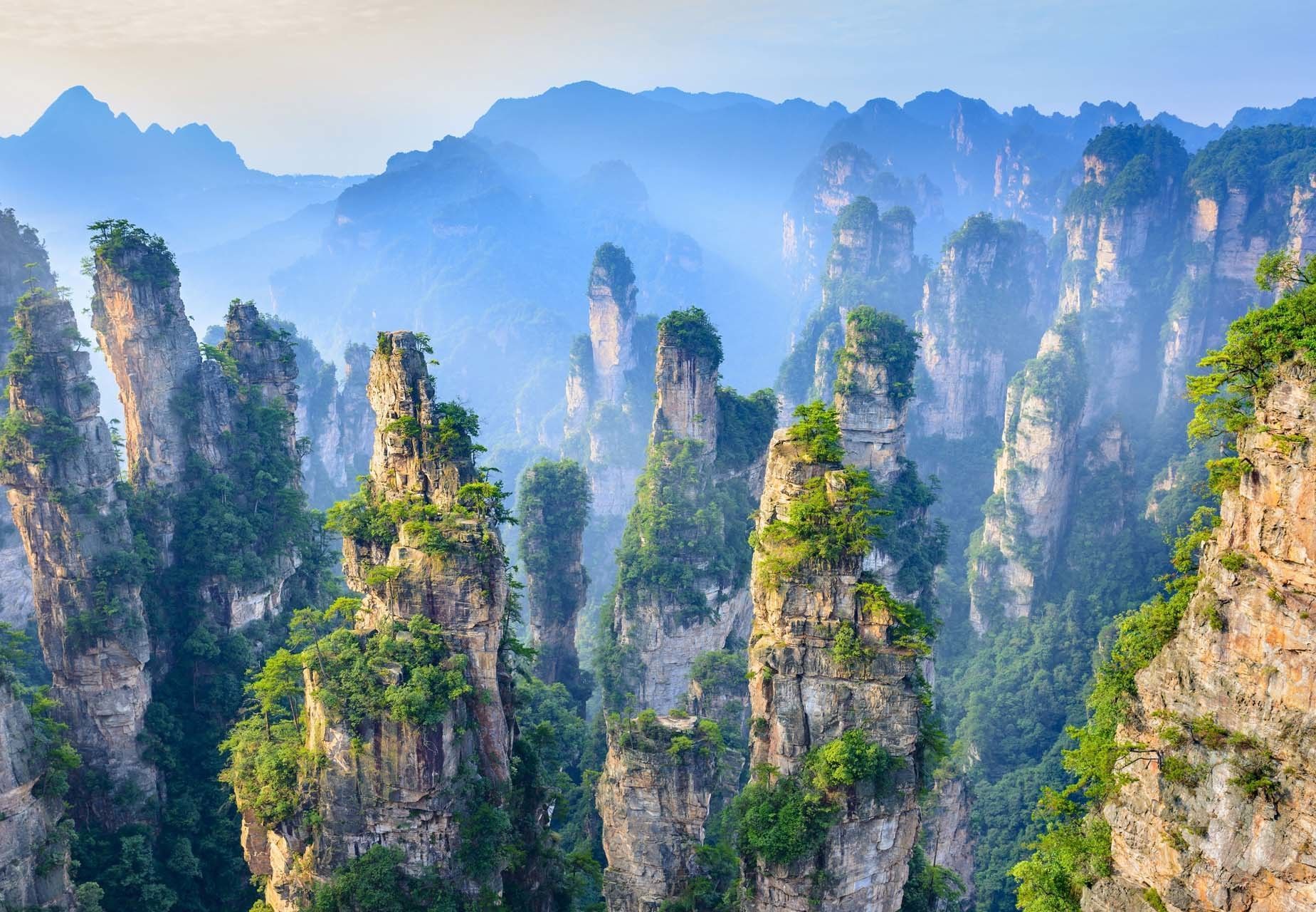 Alti e sottili pilastri di roccia sormontati da vegetazione verde si ergono da una fitta foresta, con montagne nebbiose sullo sfondo.