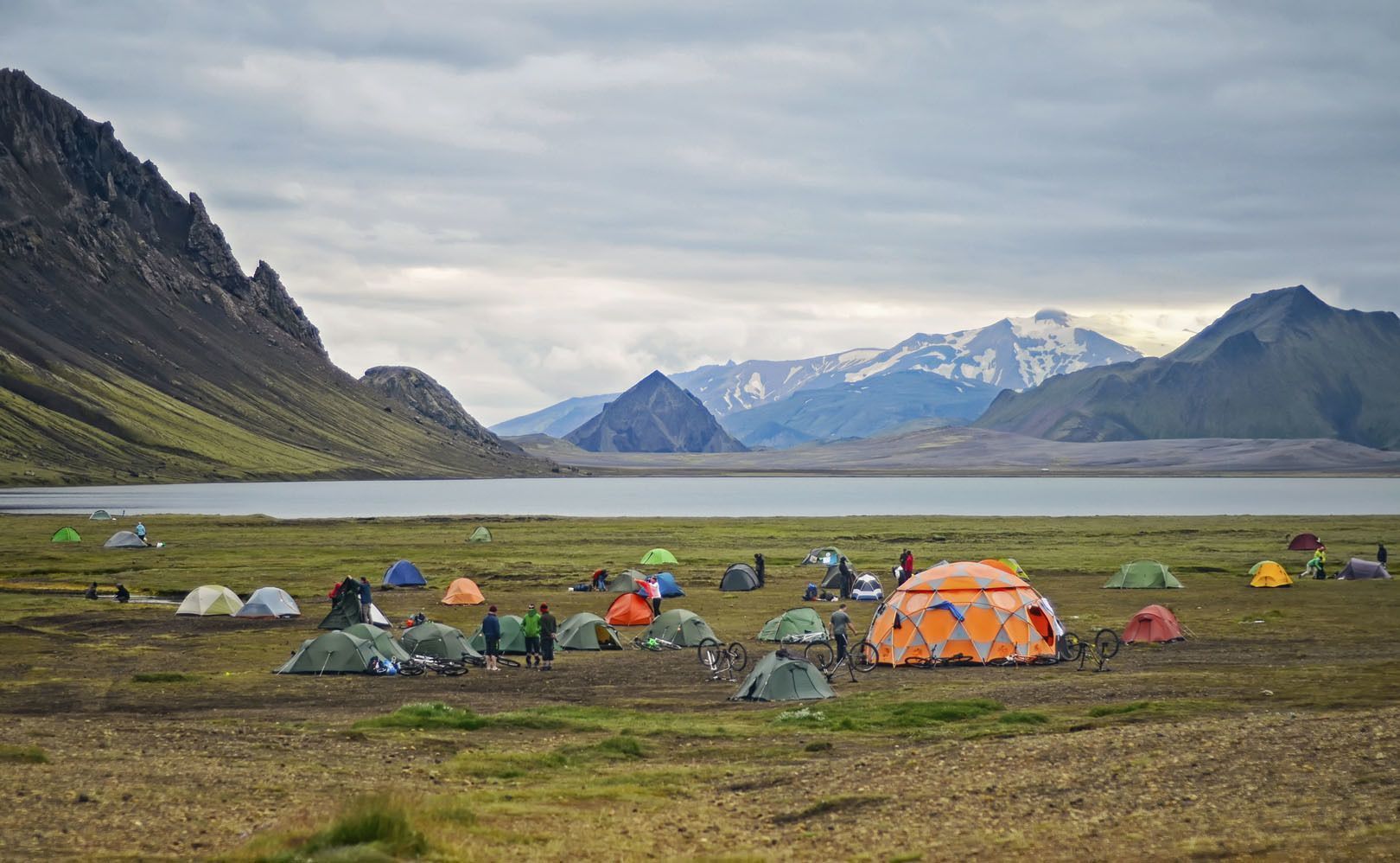 Ein großer Campingplatz mit farbenfrohen Zelten und Fahrrädern auf einer grasbewachsenen Ebene neben einem See, mit schroffen Bergen im Hintergrund unter einem bewölkten Himmel.