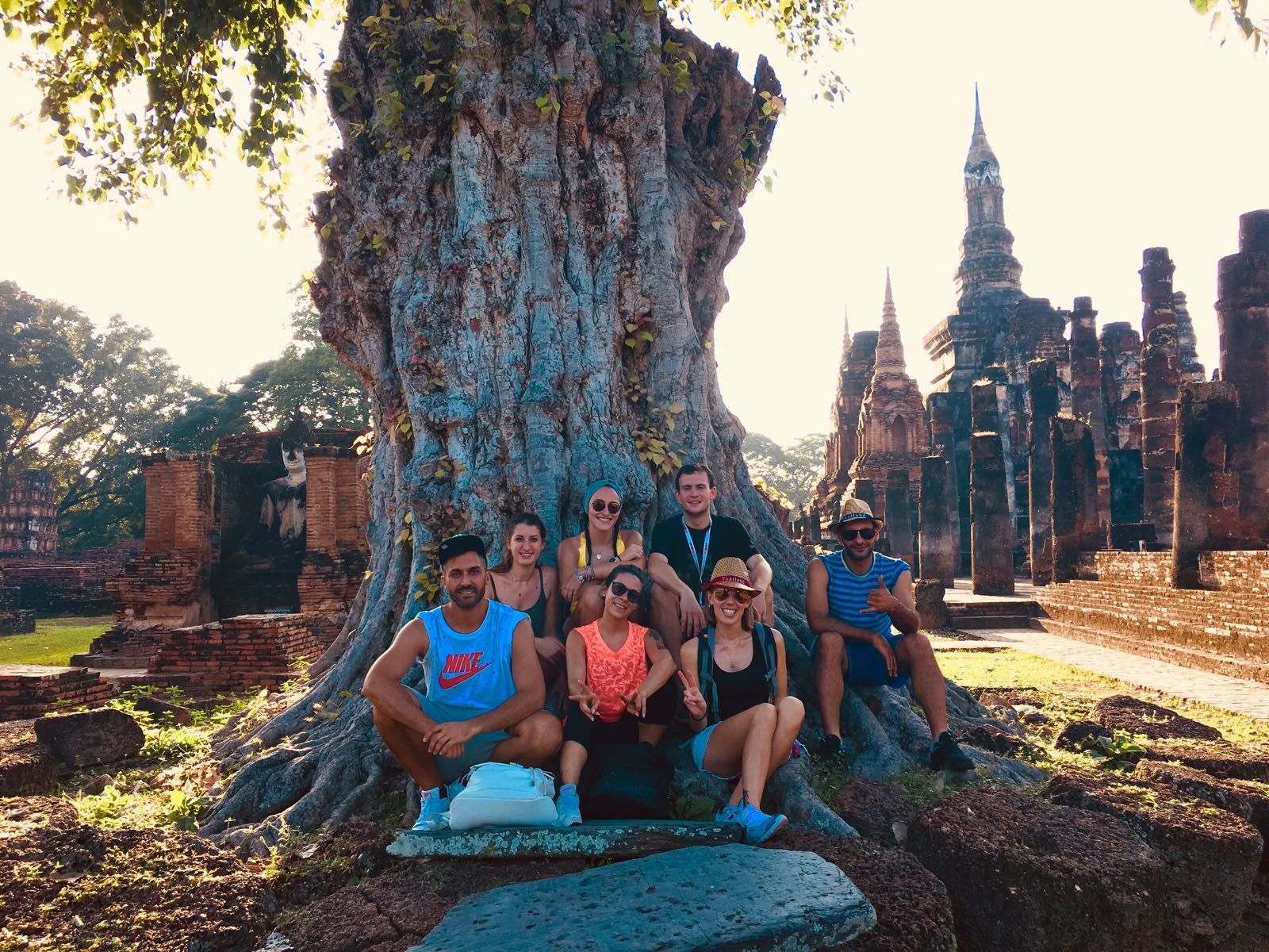 A WeRoad group trip poses for a photo at the base of a large, ancient tree with temple ruins in the background during sunset.