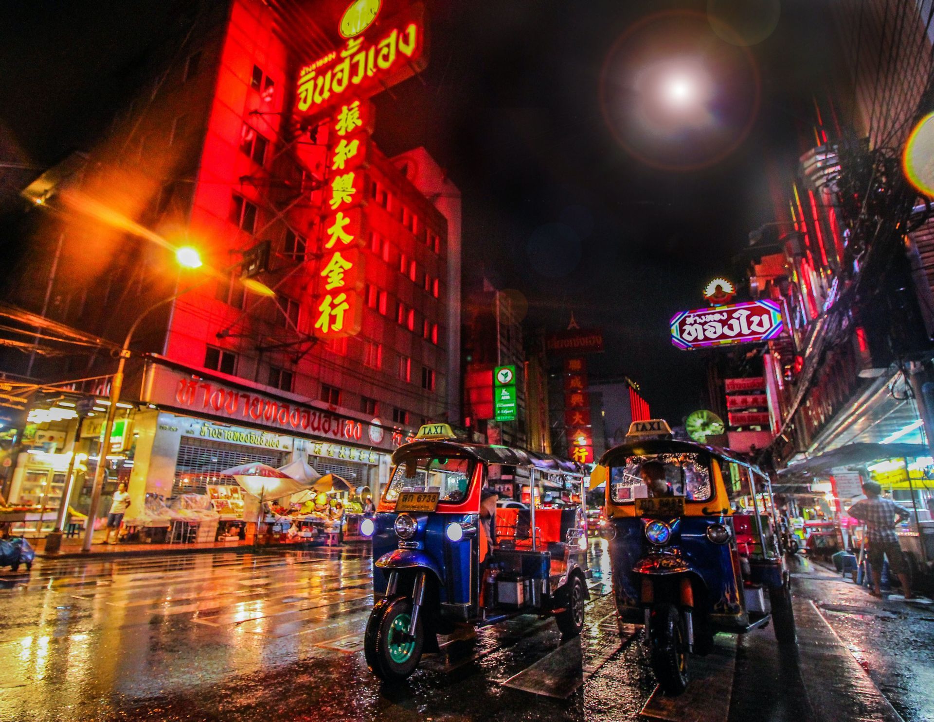 Two colorful tuk-tuks parked on a wet city street at night, illuminated by glowing red neon signs on the buildings.