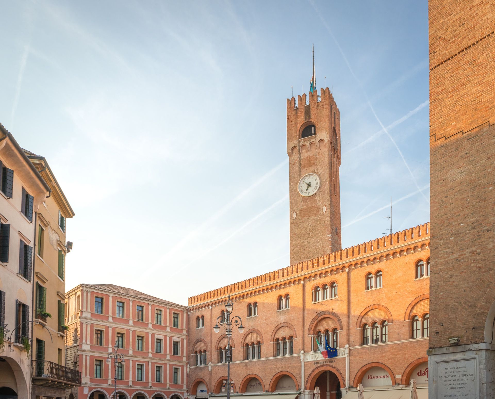 Ein sonnenbeschienener, roter Backsteinglockenturm und ein angrenzendes Bogengebäude stehen auf einem historischen Marktplatz unter einem hellblauen Himmel.