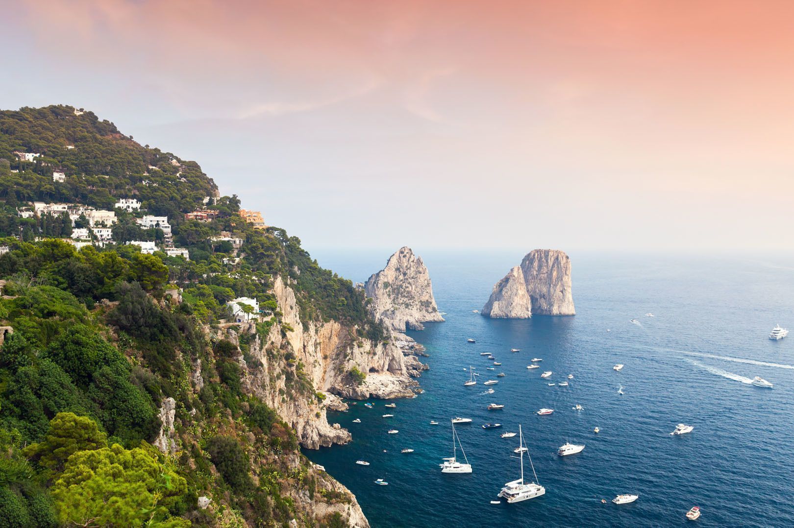 Une vue aérienne d'une côte rocheuse et verdoyante avec deux pitons rocheux et de nombreux bateaux sur l'eau sous un ciel rose et orange.