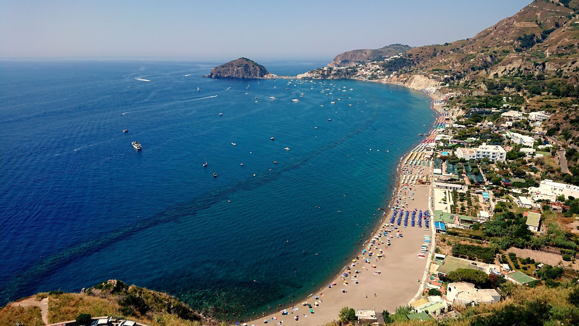 Vue aérienne d'une plage de sable bondée, avec des parasols colorés s'étendant le long d'une baie aux eaux d'un bleu profond, parsemée de bateaux et dominée par une ville côtière perchée sur une colline.