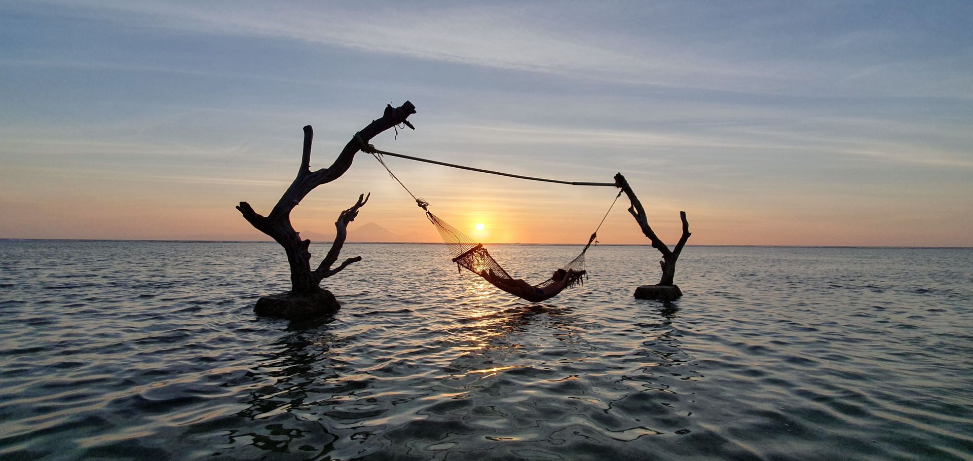 Una persona en silueta relajándose en una hamaca suspendida entre dos troncos de árboles sobre el mar al atardecer.