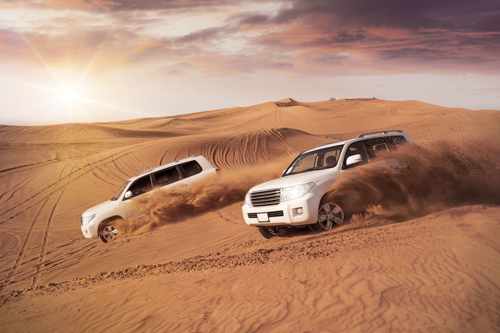 Due SUV bianchi sollevano sabbia mentre guidano su grandi dune del deserto sotto il sole al tramonto