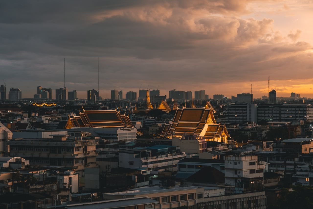 Beleuchtete Tempeldächer heben sich bei Sonnenuntergang unter bewölktem Himmel von einer dichten Stadtlandschaft mit moderner Skyline ab.
