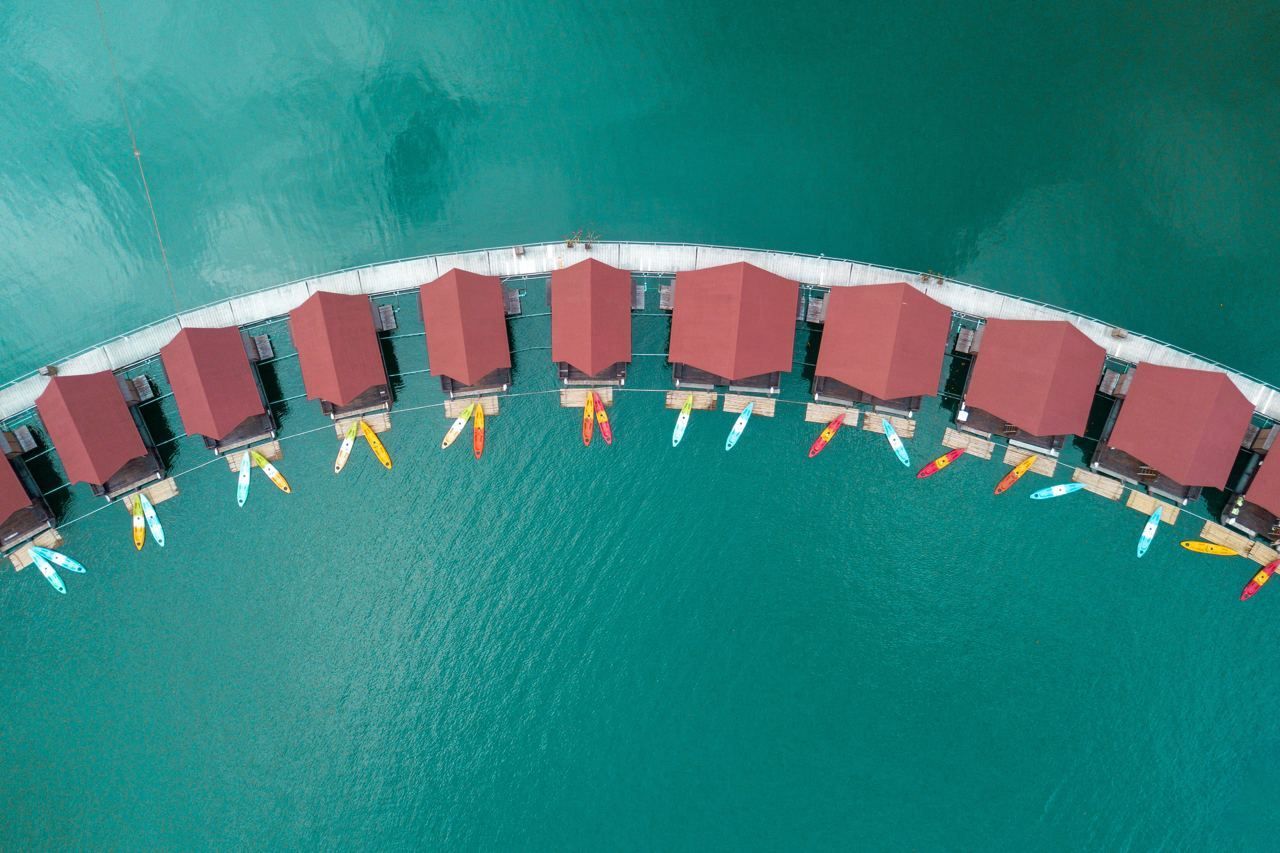Eine Luftaufnahme von geschwungenen schwimmenden Bungalows mit roten Dächern und Kajaks, die auf türkisfarbenem Wasser vertäut sind.