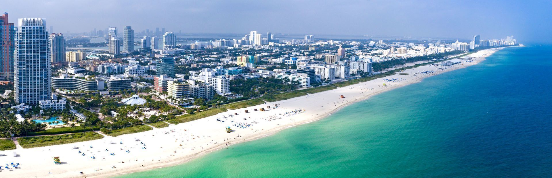 Vue aérienne d'un paysage urbain moderne avec des gratte-ciel, longeant une large plage de sable blanc et des eaux océaniques turquoise.