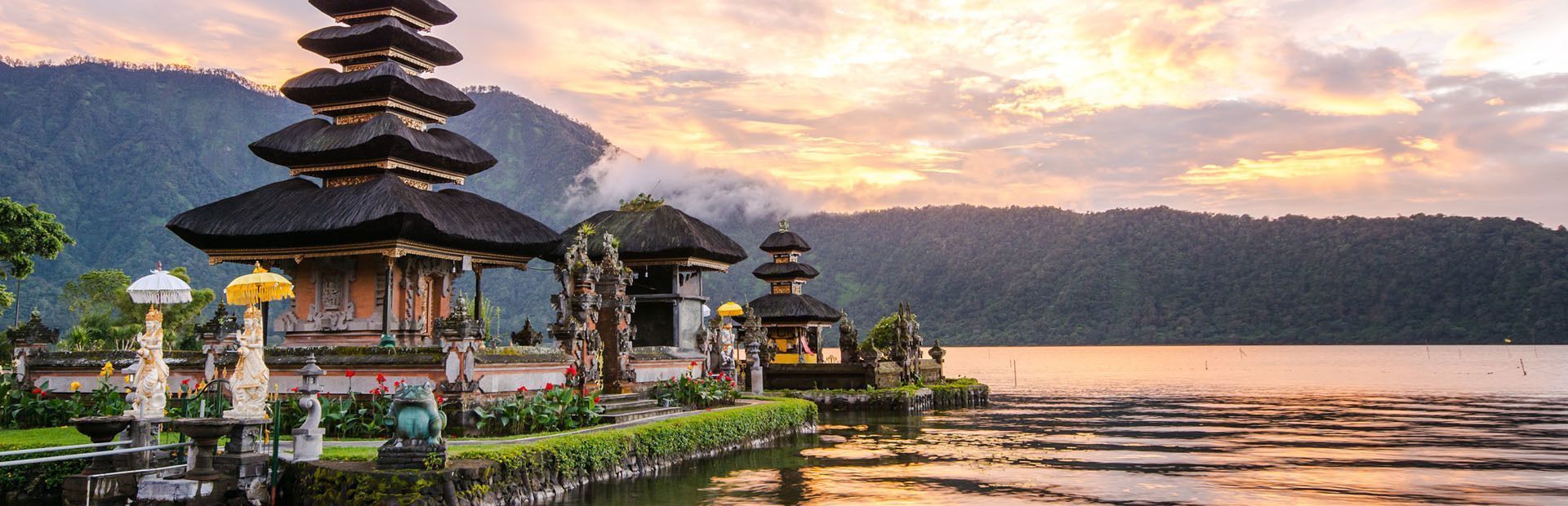 Un complexe de temples à plusieurs niveaux se trouve au bord d'un lac paisible, avec des montagnes boisées en arrière-plan sous un ciel de coucher de soleil.