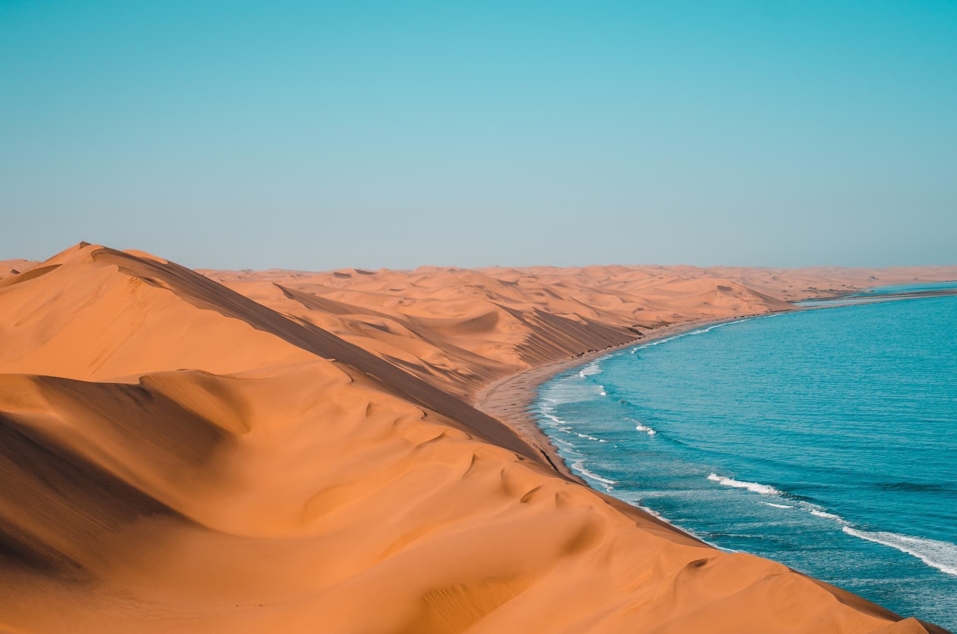 Weite orangefarbene Sanddünen treffen auf den blauen Ozean und bilden eine Küstenlinie unter klarem Himmel.