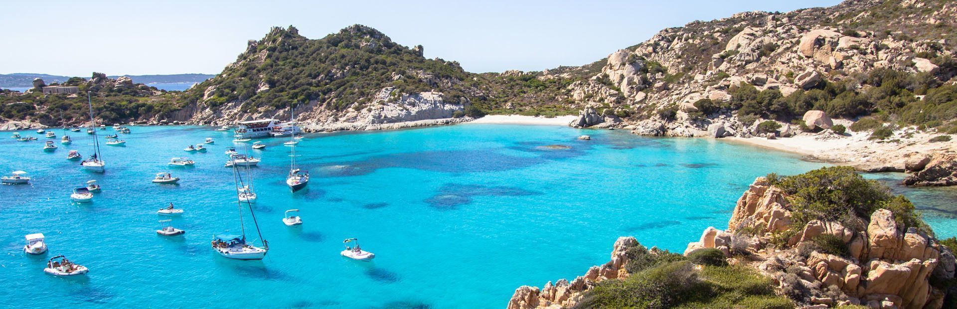 Numerose barche e barche a vela sono ormeggiate in una baia dalle acque cristalline turchesi, circondata da colline rocciose e una spiaggia sabbiosa.