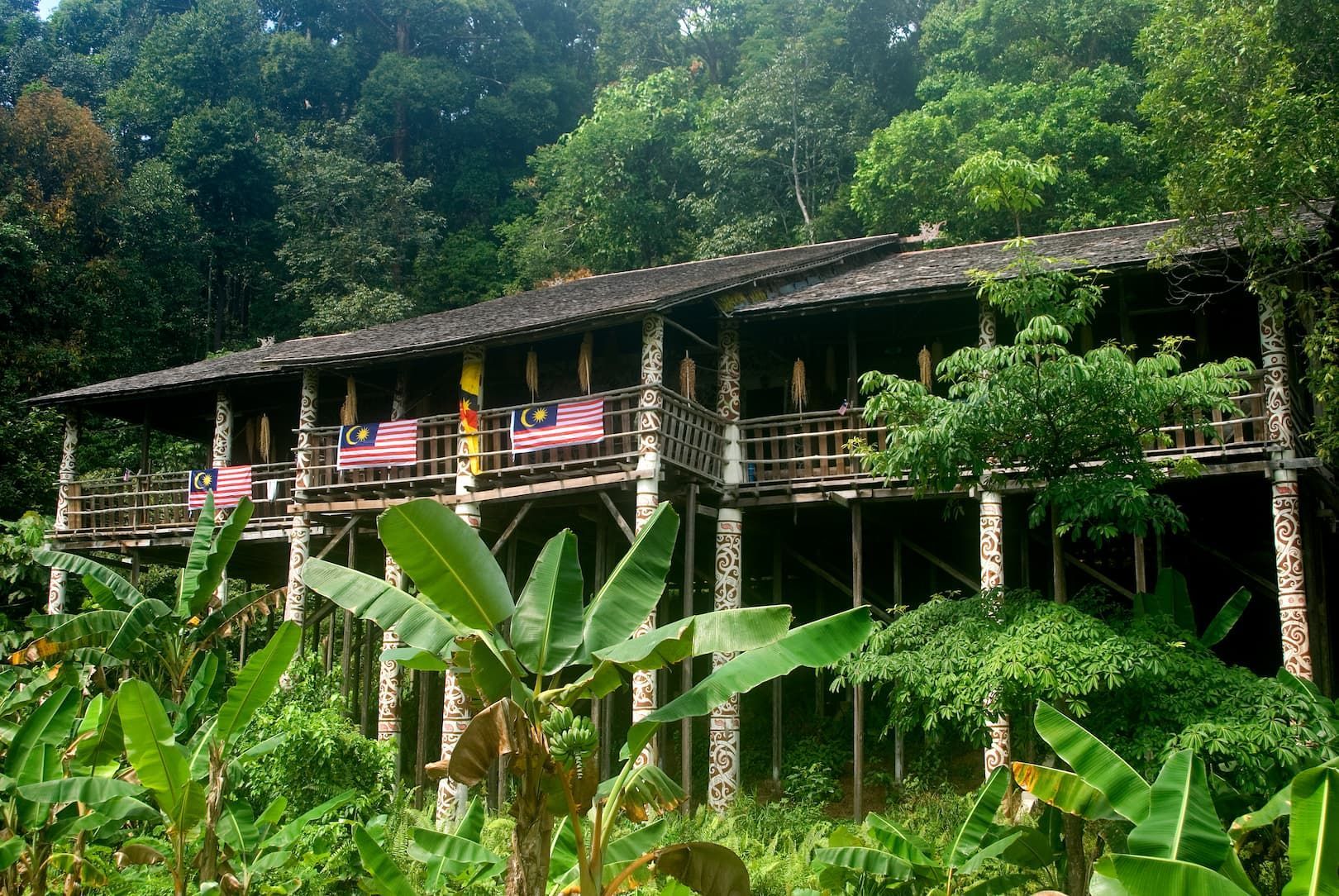 Une longhouse traditionnelle en bois, perchée sur de hauts pilotis sculptés, ornée de drapeaux malaisiens et entourée d'une végétation dense de jungle.