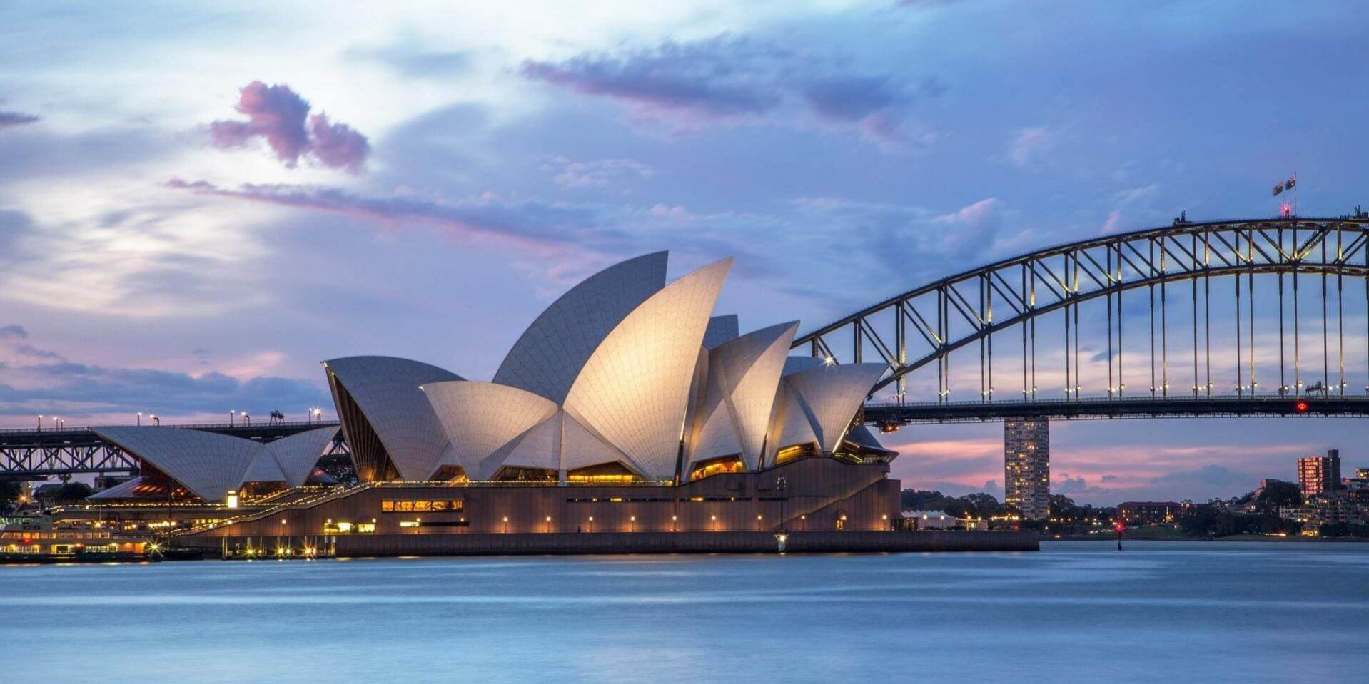 Un edificio moderno con tetti bianchi a forma di vela si erge illuminato accanto a un grande ponte ad arco sull'acqua al tramonto.