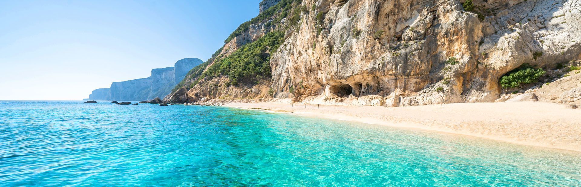 Une plage de sable isolée, baignée d'eau turquoise, au pied d'une grande falaise escarpée sous un ciel dégagé.