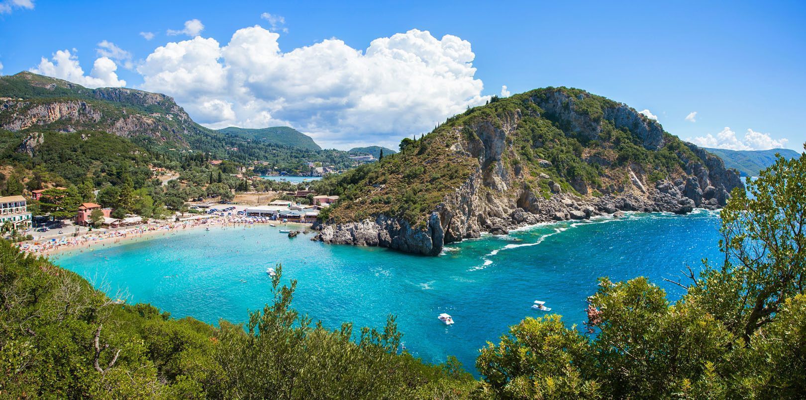 Una vista elevada de una playa concurrida con agua turquesa en una cala, rodeada de acantilados cubiertos de árboles y montañas.