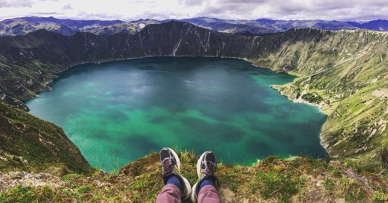 Vista in prima persona di gambe e scarponi da trekking a penzolone da un dirupo erboso, con vista su un lago vulcanico turchese.