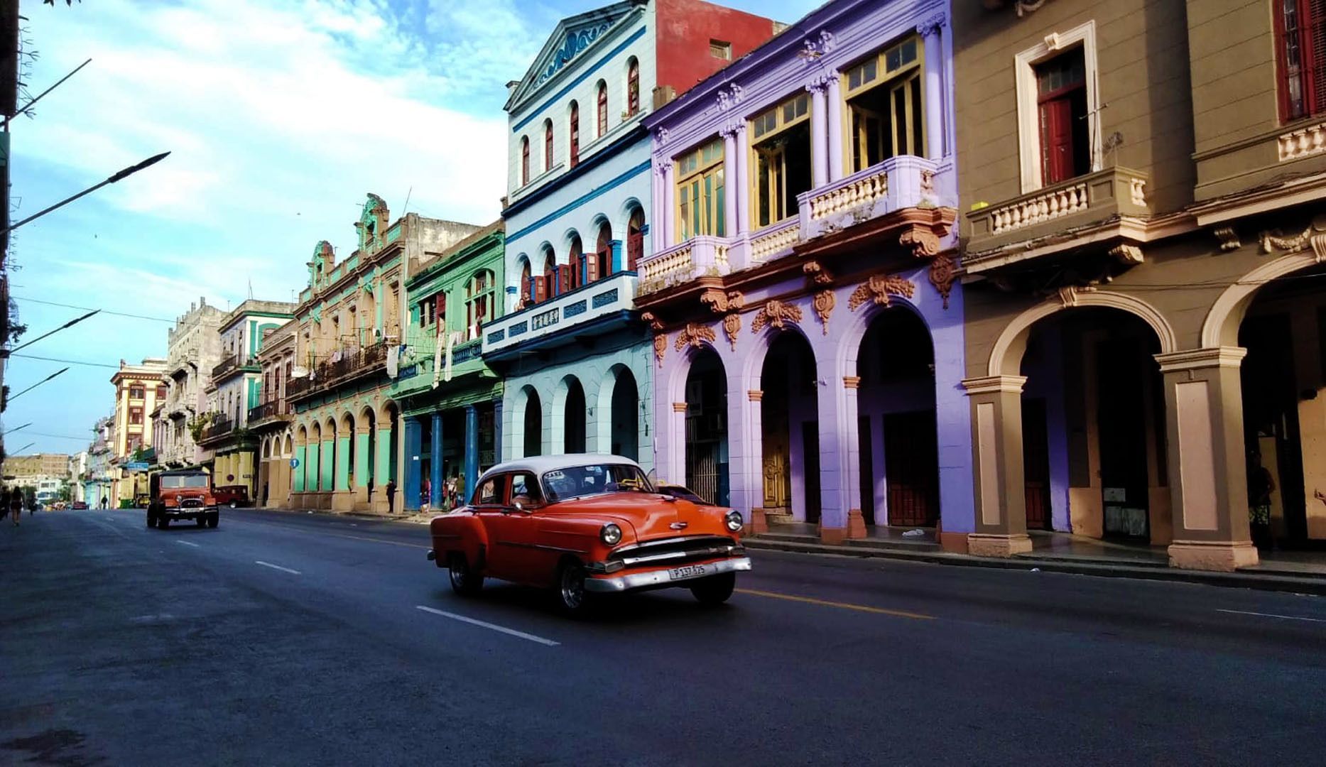 Une voiture ancienne orange roule dans une rue à côté de bâtiments coloniaux colorés avec des portes cintrées sous un ciel bleu.