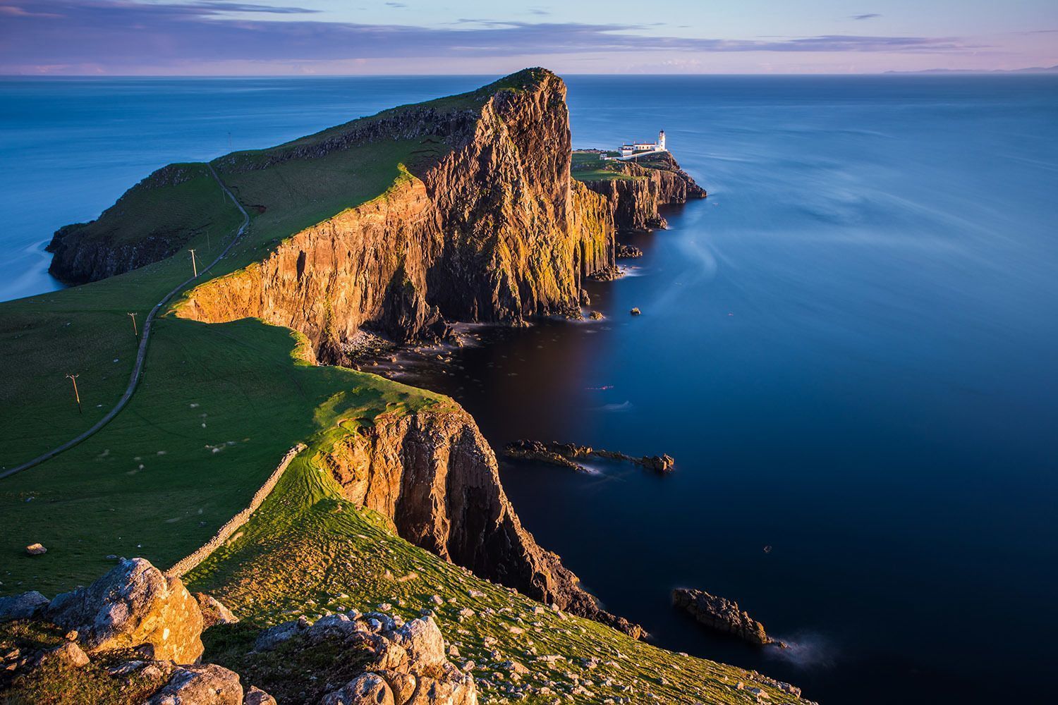 Un phare blanc se dresse sur une péninsule rocheuse et herbeuse qui s'avance dans la mer bleue et calme, baigné par la lumière chaude du coucher de soleil.