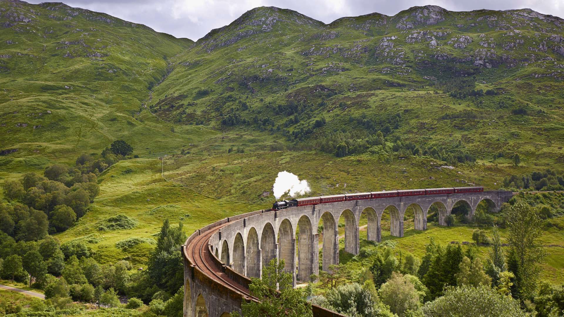 Un treno a vapore con vagoni rossi attraversa un viadotto in pietra curvo in una rigogliosa valle verde circondata da montagne.